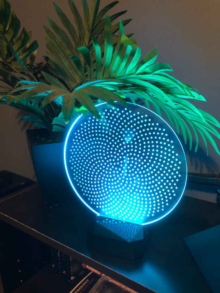 Illuminated circular acrylic phi-vortex object displayed beside foliage